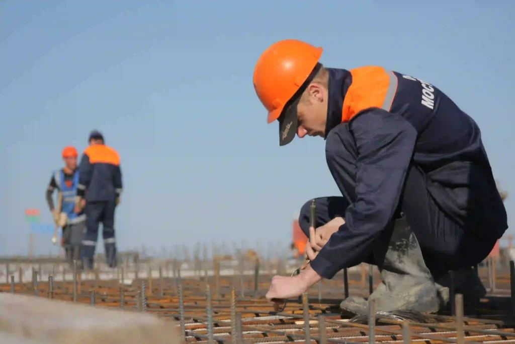 Construction worker in an orange hard hat tying steel rebar on a grid of reinforcement bars at a construction site.