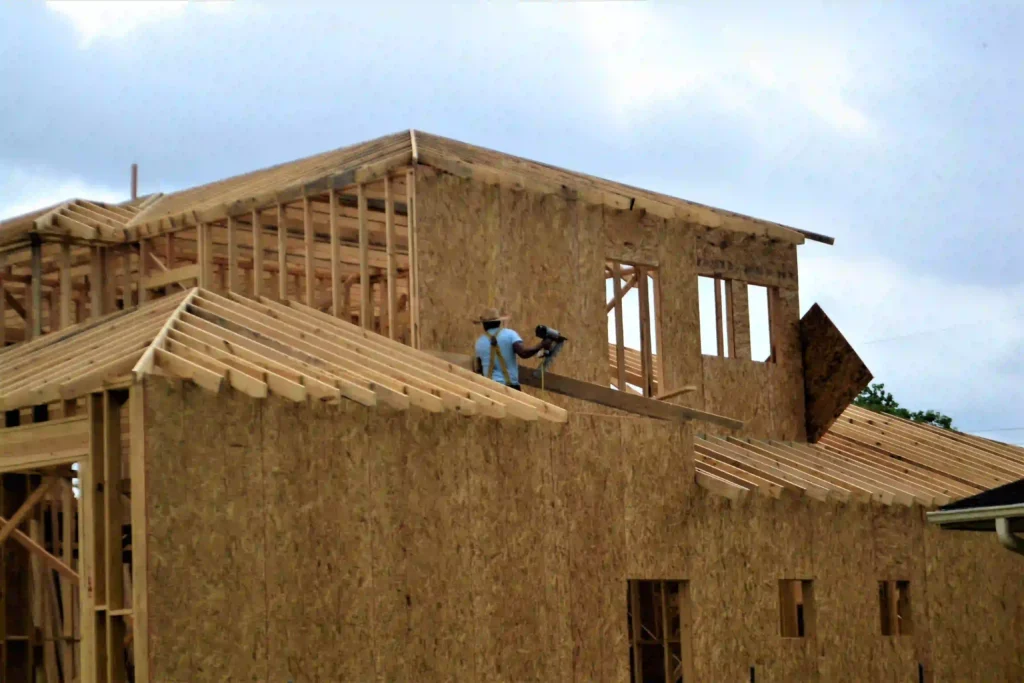 Wooden roof framing under construction on a partially built house, with a worker using a power tool on the roof structure.