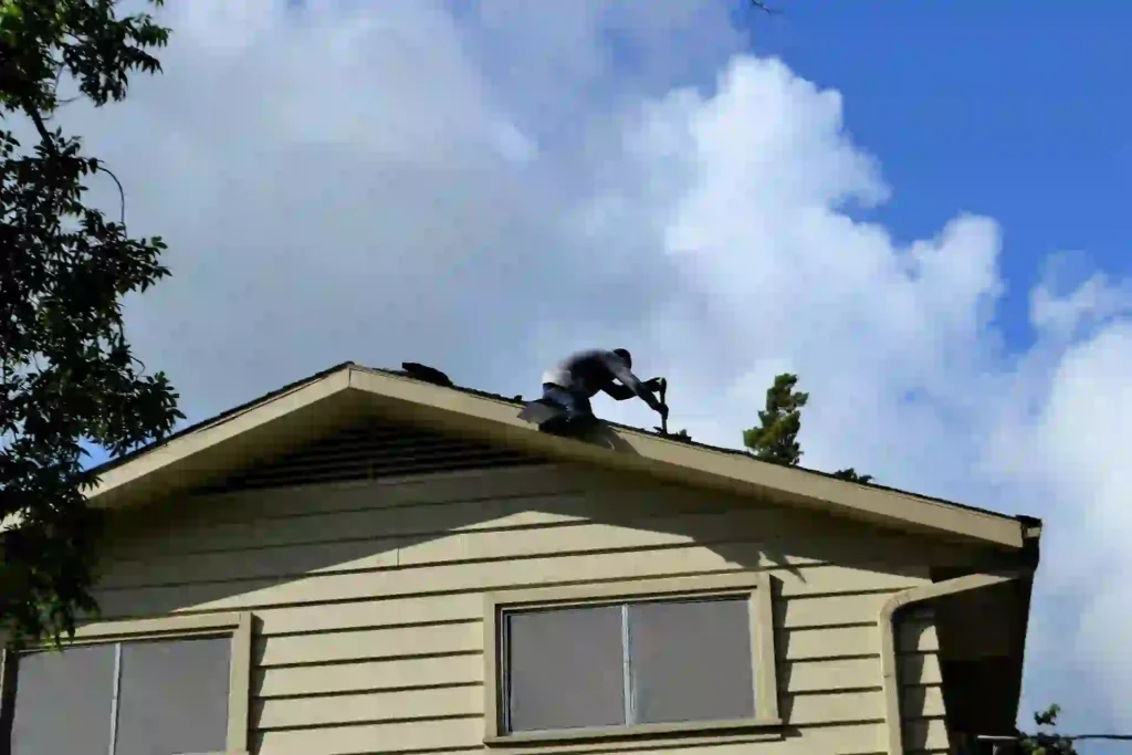 Roofing worker repairing a sloped residential roof while using a hand tool, viewed from below against a blue sky.