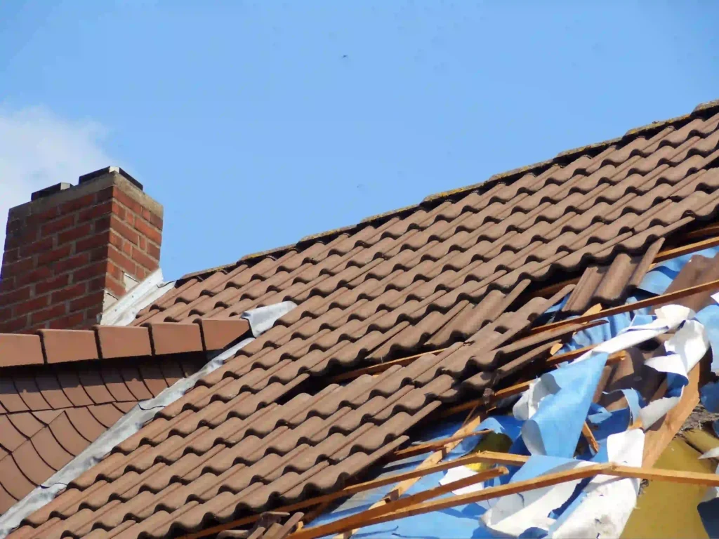 Storm-damaged clay tile roof with a brick chimney, showing broken tiles and exposed blue tarp underlayment.