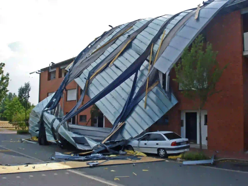 Severe storm damage showing a metal roof torn off a brick building and collapsed onto a parked car below.