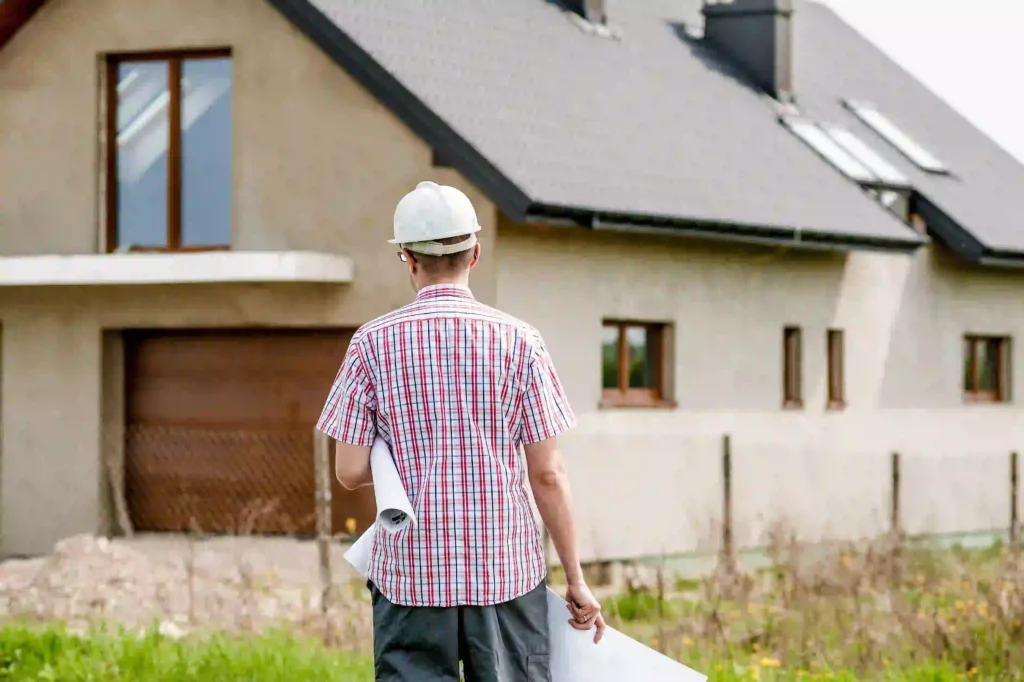 Man in a hard hat holding rolled blueprints while looking at a house with a gray shingled roof and skylights.