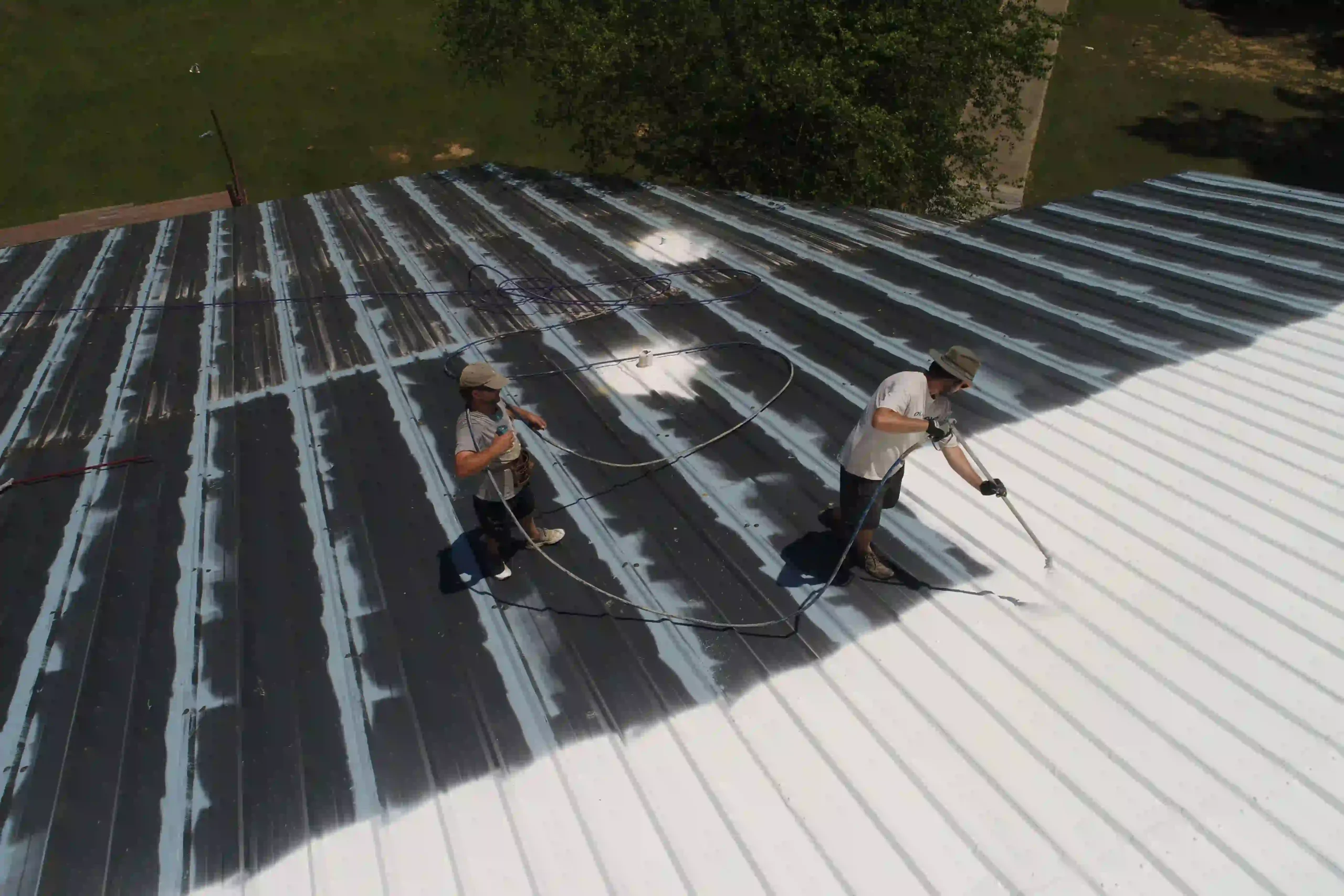 Roofing Contractor in Newton MA, Two workers applying reflective coating to a large corrugated metal roof using spray hoses.