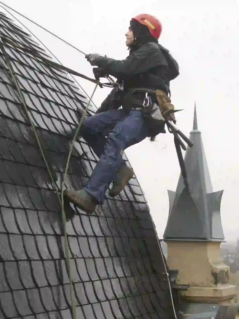 Roofing worker wearing a safety helmet and harness while working on a steep tiled roof beside a tall building tower.