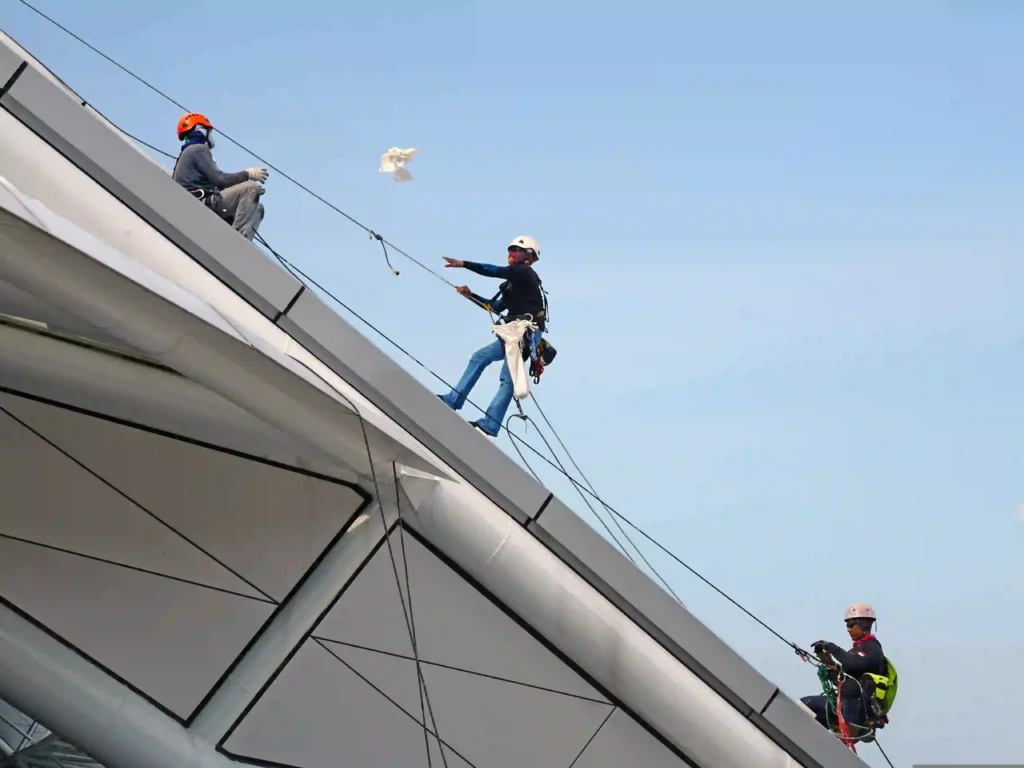 Three workers in helmets and safety harnesses rappelling down a large sloped building facade using rope access equipment.
