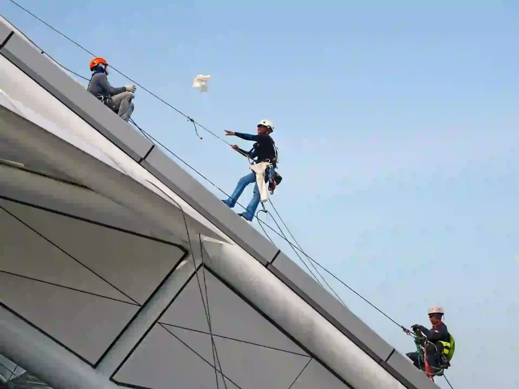 High-angle view of three maintenance technicians suspended by ropes and safety equipment while cleaning and servicing a massive metal roof arch.