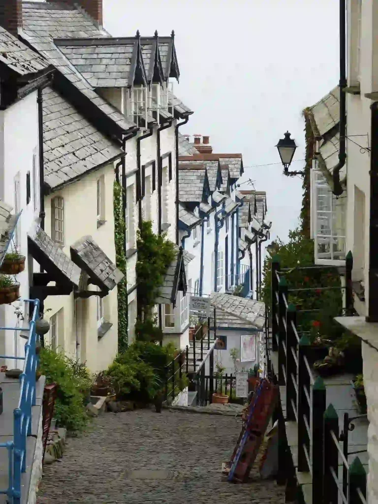 Narrow cobblestone street in a Cornwall village lined with white cottages topped with slate roofs and dormer windows.