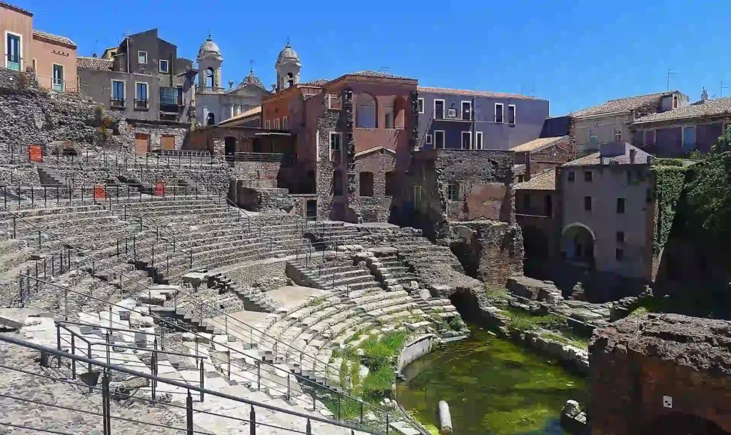 An ancient stone Roman amphitheater integrated into a historic city landscape in Catania under a clear blue sky.