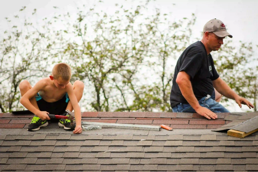 Roof Repair Estimate Newton MA, A young boy and an adult work together on an asphalt shingle roof, hammering shingles into place.