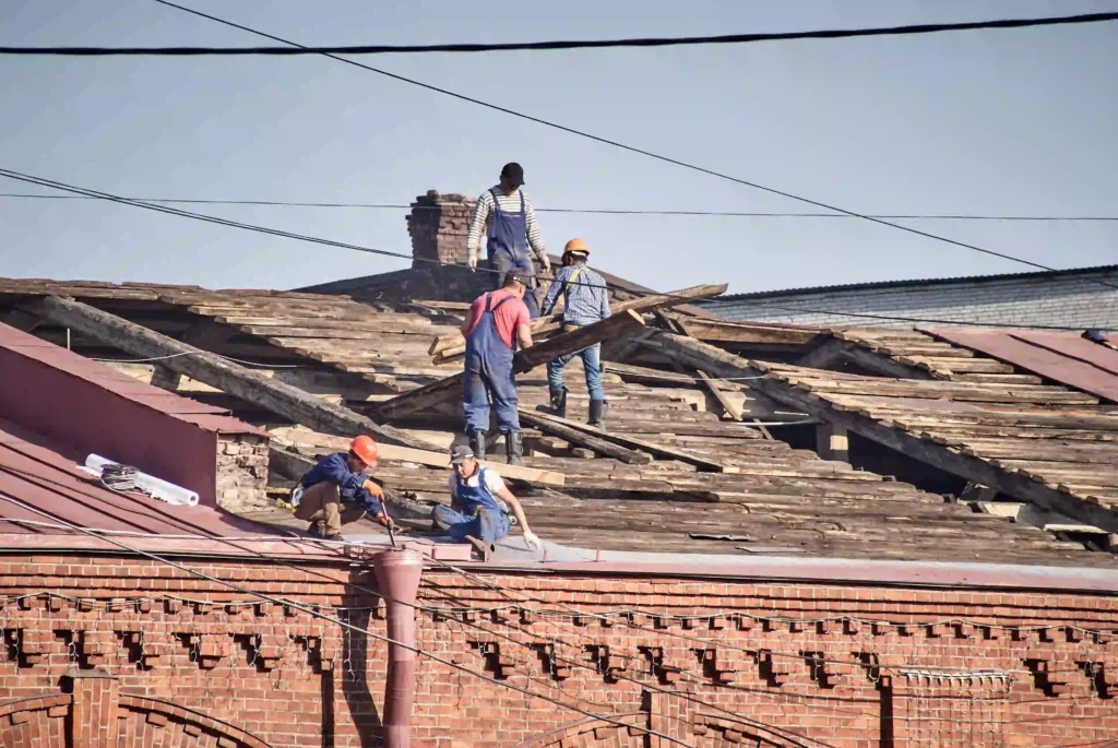 Slate Roof Repair Newton MA, Five roofers stripping an old roof down to exposed wooden rafters on a brick building with a chimney.