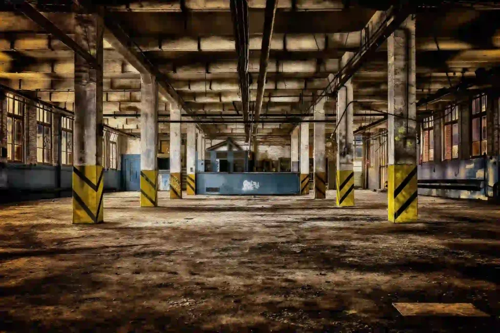 Interior of an abandoned industrial factory with concrete support columns, exposed ceiling beams, and graffiti.