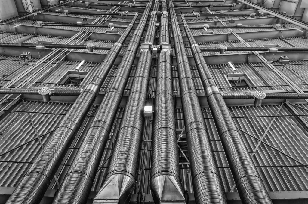 Attic Ventilation Roof Problems, Black and white view of large industrial HVAC ductwork and metal pipes running across a ceiling structure.