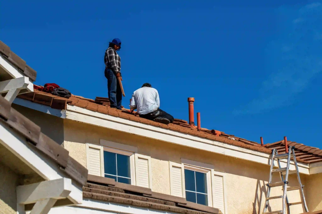 Roof Repair vs. Roof Replacement, Roofers performing residential roof installation and repairs on a sunny day using a ladder and hand tools.