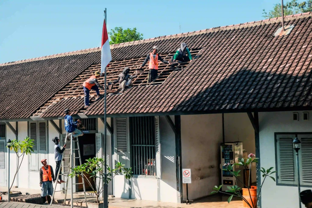 Roofing crew repairing a clay tile roof on a colonial-style building, with an Indonesian flag mounted on the facade.